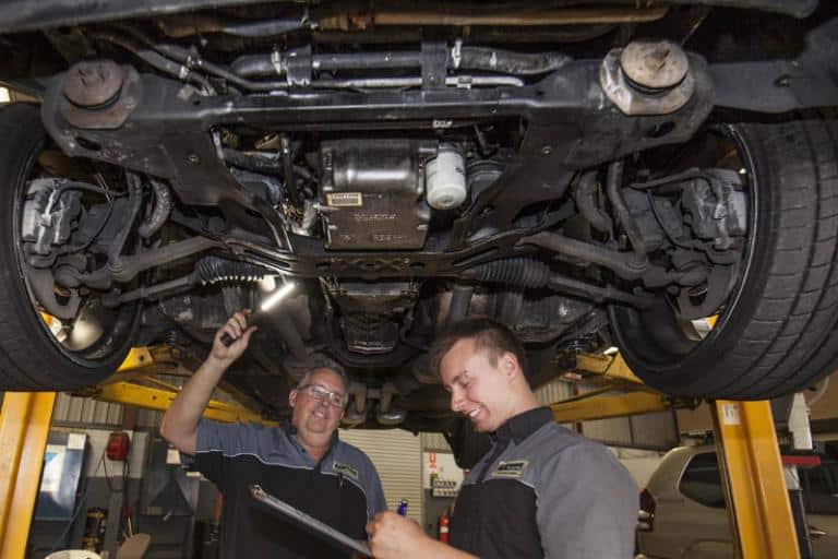 mechanics examining the underneath of a car with a torch in service centre while it is on a car lift writing down what they see